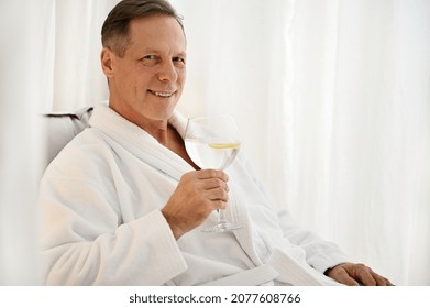 Handsome Successful Mature 50 Years Old European Man In White Terry Dressing Gown Holding Glass Of Lemon Water And Smiling Toothy Smile Looking At Camera While Relaxing In Private Room Of Wellness Spa