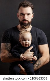 Handsome Tattooed Beared Man In Black T-shirt, Looking At Camera With Serious Face, Holding Cute Little Baby On Black Background