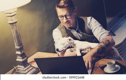 Handsome Tattooed Man In Eyeglasses Working At Home On Laptop While Sitting At The Wooden Table With Cute Dog Sleeping On His Hands.Adult Guy Using Modern Computer For Surfing Web