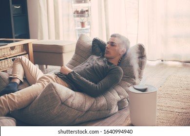 Handsome teenage guy relaxing on modern couch in living room interior. Young man resting in soft chair at home using humidifier.