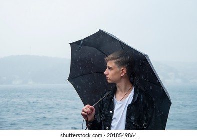 Handsome Teenager 15-18years Old, Holding An Umbrella,walks Along The Street, Natural Blue Gray Background Of The Sea.Close Up Emotional Portrait Of Caucasian Young Man Under Rain.Selective Soft Focus