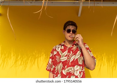 Handsome Thai Man Wearing Red Hawaii Shirt And Sunglasses, Licking Loliipop Isolated Over Yellow Background. Summer Time Concept.