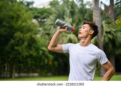 Handsome, Thirsty And Sweaty Millennial Asian Athletic Man In Sportswear Drinking Water Or Protein Drink From Bottle After Training In The Park.