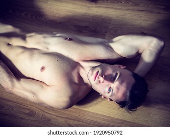 Handsome totally naked muscular young man laying down on hardwood floor at home in seductive attitude, looking at camera