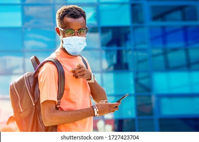 Handsome Trendy African Americans Nerd Male With Glasses In Stylish Clothes Colorful T-shirt With A Backpack On The Shoulders Stand Against The Background Of The Blue Windows Of The University