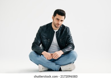 Handsome Trendy Man Sitting On Floor Isolated On White Studio Background. Men's Beauty Concept. Young Male Wearing Jeans, White T-shirt And Black Leather Jacket Posing Sitting With Legs Crossed