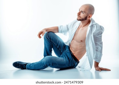 handsome Turkish man with bald head sits on floor against a white wall, his open shirt revealing his well-defined muscular abs. He look thoughtfully towards left upper corner - Powered by Shutterstock