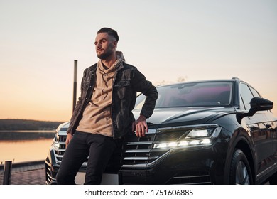 Handsome unshaved man in fashionable clothesleaning on his his black car.