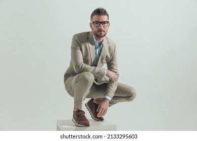 Handsome Unshaved Man In His 40s With Elbow On Knee Crouching And Squatting While Looking Up Dressed In A Brown Suit With Open Collar Shirt