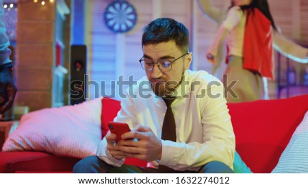 Handsome unsociable guy using social media or browsing news on smartphone getting upset on night party. People in background having fun celebration.