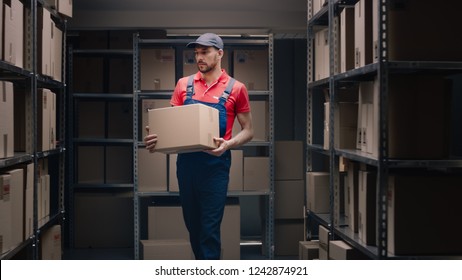 Handsome Warehouse Worker Walks In Storeroom With A Cardboard Box.