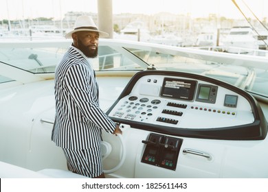 A Handsome Wealthy Mature Bearded Black Guy In A Hat And A Striped Summer Costume With Shorts Is Driving His Luxury Yacht To Away From The Berth, Turning The Steering Wheel And Looking At The Camera