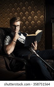 Handsome Well-dressed Man Sitting By The Fireplace In A Room With Classic Vintage Interior And Reading A Book. Fashion. Luxury.