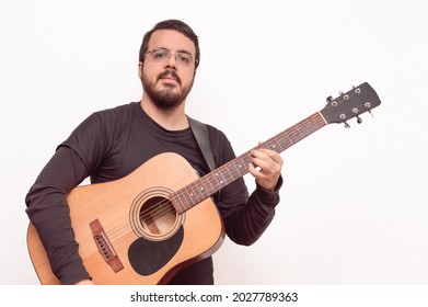 Handsome White Hispanic Latin Adult Male With Short Beard, Short Hair, Black Dress And Lens Posing While Playing A Chord With An Acoustic Guitar On A White Background.