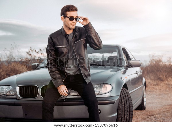handsome white man on luxury car wearing sunglasses and\
leather jacket. A stylish male model with status looking out on\
beautiful sunset. 