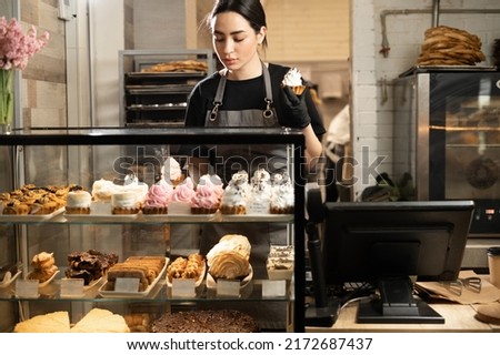 Handsome woman coffee shop employee placing pastry and cake in bakery refrigerator showcase at cafe. Small business, part time job concept