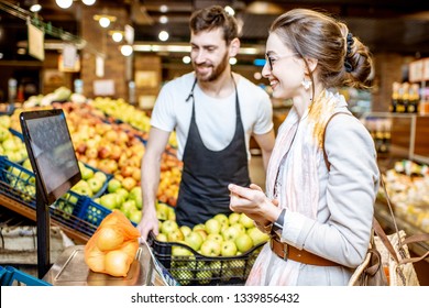 Handsome worker in uniform helping young woman customer to weigh apples on the scales in the supermarket