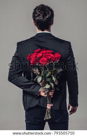 Handsome yound man in suit is standing with red roses behind the back on grey background.