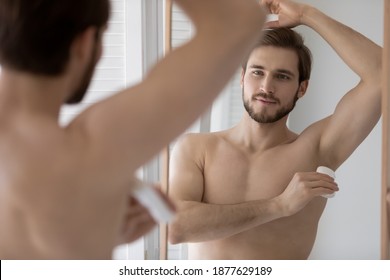 Handsome Young 30s Shirtless Man Applying Deodorant In Arm Pit, Looking In Mirror. Happy Millennial Man Preventing Sweating, Using Underarm Antiperspirant After Morning Showering Hygienic Routine.