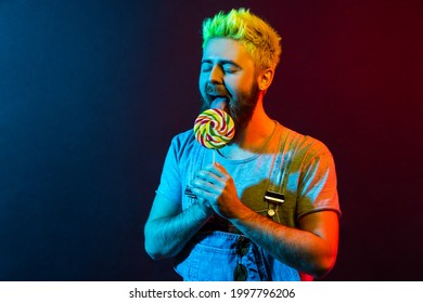 Handsome Young Adult Bearded Hipster Man With Lolly Pop In Hands, Enjoying Licking Tasty Candy, Keeping Eyes Closed, Wearing Denim Overalls And T Shirt. Colorful Neon Light, Indoor Studio Shot.