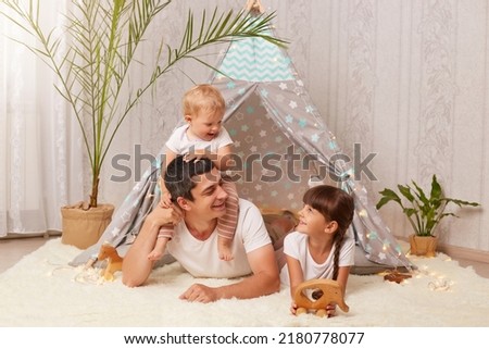 handsome young adult father with little daughters having fun and playing in peetee tent in children room at home, infant baby sitting on dad's back, family with happy expressions.