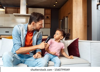 Handsome Young Adult Father Sitting On Comfort Sofa With His Afro American Daughter, Holding Microphone In Hands, Taking Interview Of Singing And Spending Free Time With Offspring