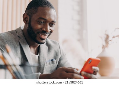 Handsome Young African American Businessman Using Mobile, Chatting Online While Sitting At Office Desk, Typing Message And Looking At Phone Screen With Positive Smile, Distracted From Work