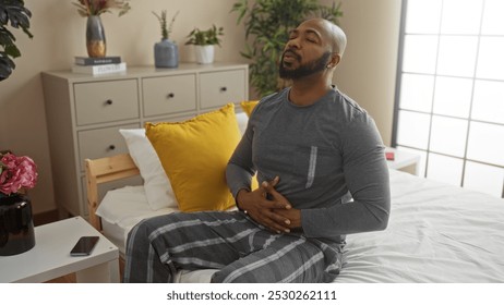 Handsome young african american man sitting on bed in bedroom holding stomach in indoor home setting