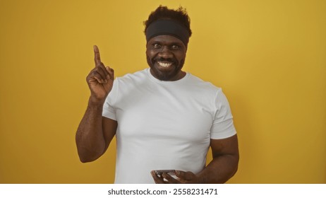 Handsome, young, african american man with a beard wearing a white t-shirt and headband, smiling and pointing up while standing against an isolated yellow background. - Powered by Shutterstock