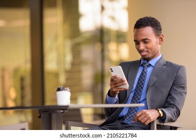 Handsome young African businessman wearing suit in city and sitting at coffee shop - Shutterstock ID 2105221784