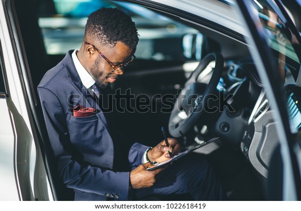 Handsome young african car salesman standing at\
the dealership holding a\
tablet
