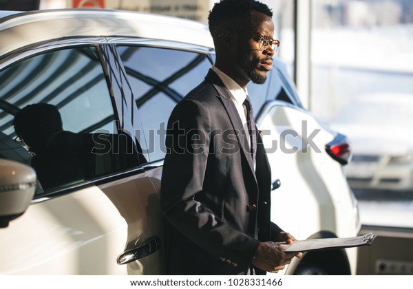 Handsome young african car salesman standing at\
the dealership holding a\
tablet