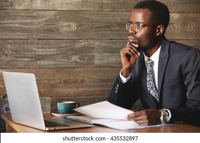 Handsome young African man wearing formal suit sitting at a coffee shop with pensive look, thinking of business plans, holding a pen, leaning his elbow on the table, signing papers, working on laptop