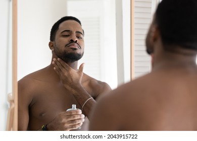 Handsome young African shirtless man reflected in mirror applying to neck perfume or to skin alcohol-free aftershave lotion. After shaving skincare product ad, personal hygiene morning routine concept