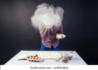 Handsome And Young Afro African Man Preparing Homemade Cakes American Pie From Fresh Dough Hands Dirty By Flour, On The Table Are Eggs, Rolling Pin And Recipe Book On A Black Background In The Studio