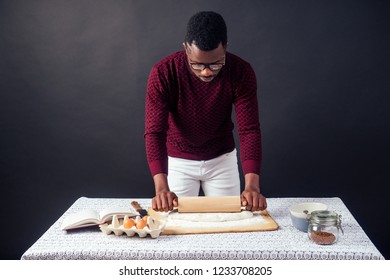 Handsome And Young Afro African Man Preparing Homemade Cakes American Pie From Fresh Dough Hands Dirty By Flour, On The Table Are Eggs, Rolling Pin And Recipe Book On A Black Background In The Studio