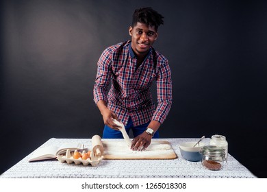 Handsome And Young Afro African Man Preparing Homemade Cakes American Pie From Fresh Dough Hands Dirty By Flour, On The Table Are Eggs, Rolling Pin And Recipe Book On A Black Background In The Studio