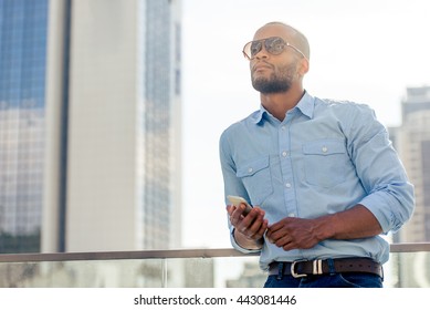 Handsome young Afro American businessman in sun glasses is holding a smartphone, looking away and thinking while standing outdoors - Powered by Shutterstock