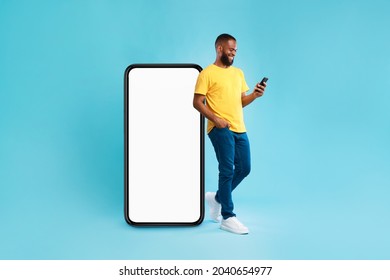 Handsome young Afro man using mobile phone while standing near huge smartphone with empty screen over blue studio background. Mockup for website or application, space for advertisement