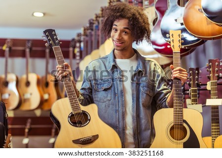 Handsome young Afro-American man holding two guitars in both hands and smiling while standing in a musical shop