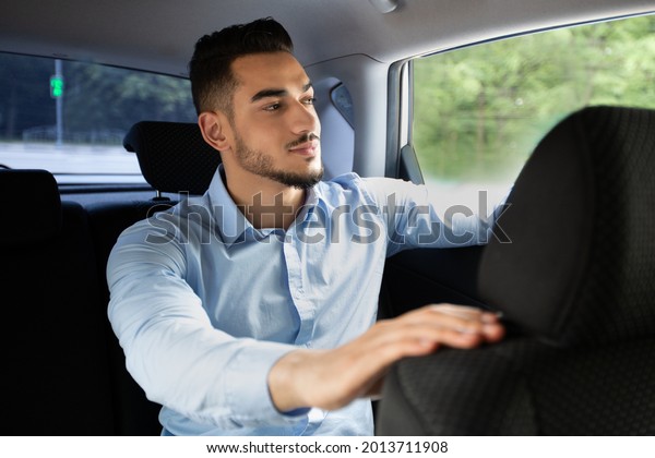 Handsome young\
arab businessman in nice stylish outfit going to office by taxi or\
corporate car with driver, sitting at back seat and looking through\
window, smiling, closeup\
portrait