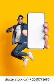 Handsome young Arab guy jumping on air, pointing at cellphone with empty screen on orange studio background. Millennial Muslim man demonstrating mockup for mobile app design - Shutterstock ID 2060771696