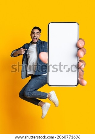 Handsome young Arab guy jumping on air, pointing at cellphone with empty screen on orange studio background. Millennial Muslim man demonstrating mockup for mobile app design