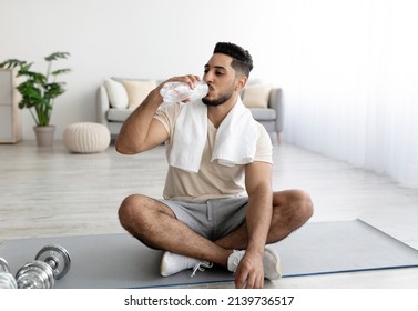 Handsome Young Arab Guy Sitting Cross Legged On Yoga Mat, Drinking Water After Domestic Training Indoors. Attractive Millennial Middle Eastern Man Keeping Hydrated During Home Workout