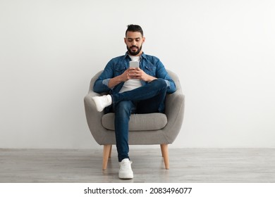 Handsome young Arab guy using mobile phone, chatting online, sitting in armchair, surfing social media against white studio wall. Distance communication, blogging concept