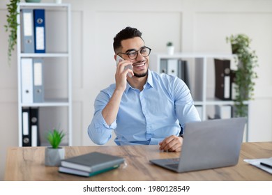 Handsome young Arab man in formal wear talking on smartphone, enjoying conversation at workplace. Attractive millennial entrepreneur speaking with customer while working on laptop at office