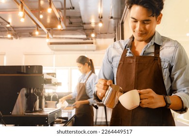 Handsome young asian barista pours milk froth into latte art coffee cup to decorate it beautifully appetizing mellow together with beautiful barista staff serving customers in the coffee cafe. - Shutterstock ID 2264449517