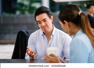 A Handsome And Young Asian Business Man In A Crisp White Shirt Is Talking To A Woman In A Suit. He Is Making A Reluctant, Grudging And Unimpressed Expression On His Face As He Thinks Something Over.