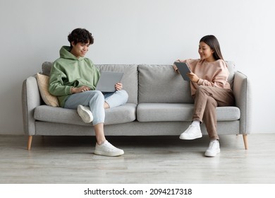 Handsome young Asian guy and his girlfriend using modern gadgets while sitting on couch at home, copy space. Full length of millennial couple communicating online via laptop and tablet pc