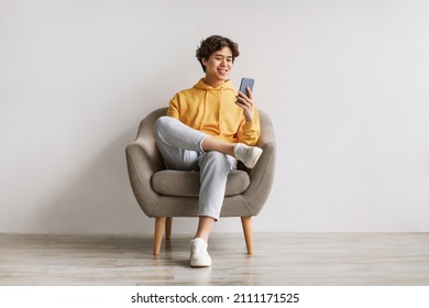 Un joven y guapo asiático usando teléfonos celulares, internet de surf o medios sociales, sentado en un sillón, disfrutando de las tecnologías contemporáneas, revisando la nueva aplicación móvil contra la pared de un estudio blanco